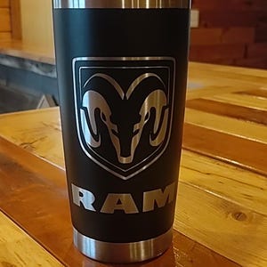 May include: Black insulated tumbler with a silver rim and base. The tumbler features the RAM logo in silver, a shield with two ram heads, and the word "RAM" below. The tumbler is on a wooden surface.
