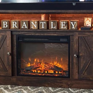 May include: A dark brown wooden media console featuring an electric fireplace insert with glowing artificial logs. The upper shelf displays eight wooden blocks spelling BRANTLEY, a circular decorative plate, a small framed photo, and a green football.