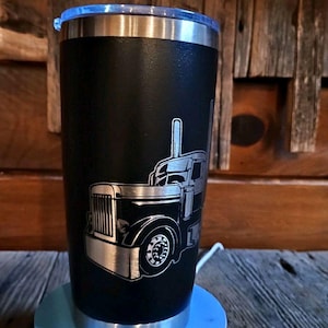 May include: Black stainless steel tumbler with a silver truck design. The tumbler has a clear plastic lid and a silver rim. The truck design is etched into the black surface, showcasing the details of the vehicle.