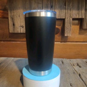 May include: A black insulated tumbler with a silver rim and a clear plastic lid. The tumbler is cylindrical and stands on a light blue and white base. The background is a wooden wall.
