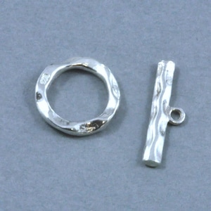 May include: A silver toggle clasp set consisting of a circular ring and a T-bar component. Both pieces feature a textured, hammered surface finish. The ring is stamped with a small hallmark, and the T-bar includes a small loop for attachment.