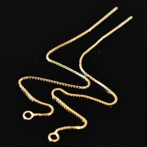 Op de afbeelding: Een delicate goudkleurige ketting met een box chain design. De ketting heeft twee dunne, rechte staven aan de bovenkant en twee kleine lussen aan de onderkant. De ketting wordt gepresenteerd tegen een zwarte achtergrond.