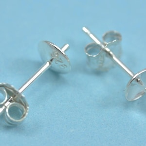 May include: Two silver earring posts with butterfly backs. The posts have a flat, round base and a slender post for insertion. The butterfly backs are designed to secure the earrings. The background is a solid light blue.
