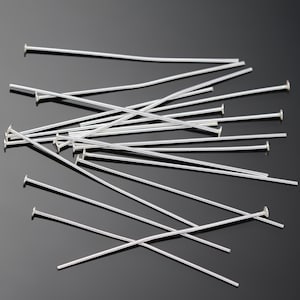 Op de afbeelding: Een stapel zilverkleurige kopspelden, ook wel bekend als sieradenspelden, met platte koppen. De spelden liggen verspreid op een zwart oppervlak.