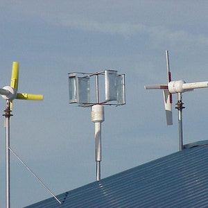 Puede incluir: Tres tipos diferentes de turbinas eólicas montadas en un techo de metal. Las turbinas están hechas de metal y plástico y tienen diferentes diseños de palas. Las turbinas están todas orientadas en la misma dirección.