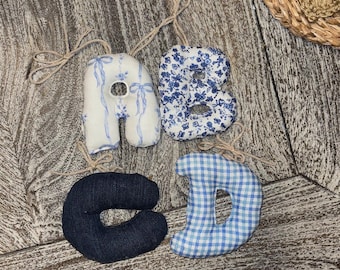 May include: Four fabric letters: A, B, C, and D. The letter A is white with blue bows, B is blue floral, C is dark blue denim, and D is blue and white gingham. Each letter has a twine loop for hanging.