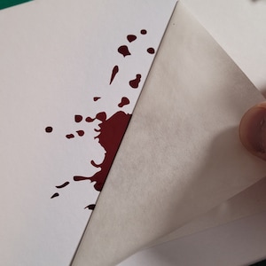 May include: A white sheet of paper with a dark red splatter design. A corner of the paper is being lifted, revealing the blank side. The image is on a green cutting mat.