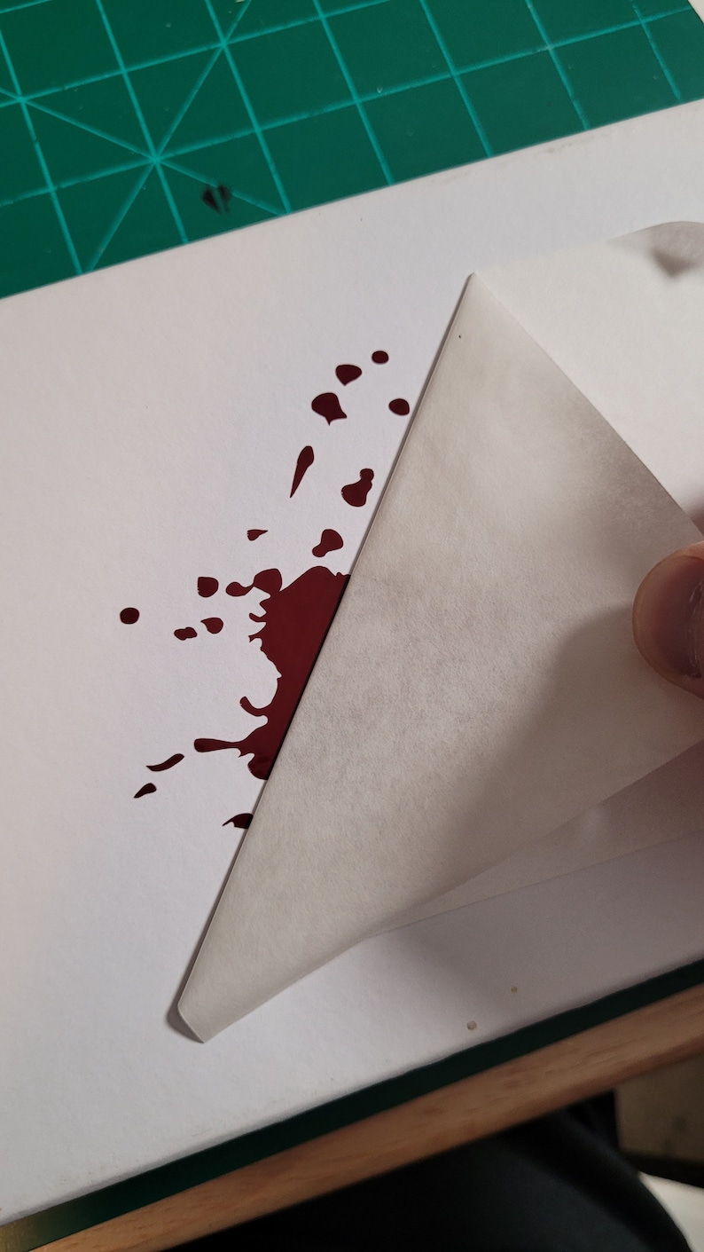 May include: A white sheet of paper with a dark red splatter design. A corner of the paper is being lifted, revealing the blank side. The image is on a green cutting mat.