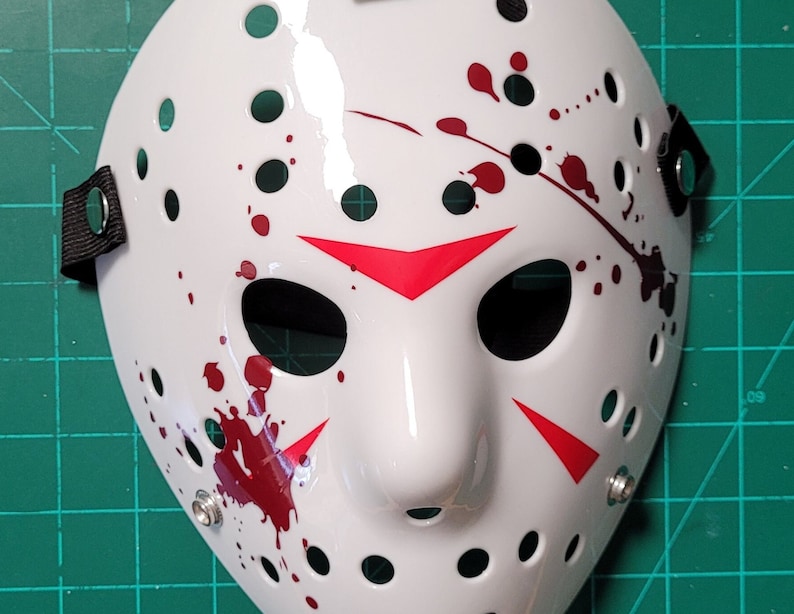 May include: White hockey mask with black eye holes and numerous circular ventilation holes. The mask features red accents and blood splatter details. Black straps are attached on each side for securing the mask.