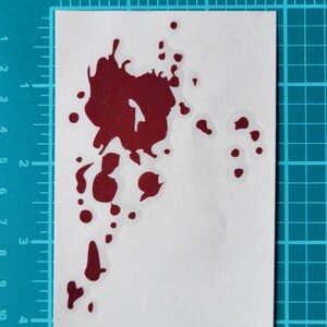 May include: A red blood splatter sticker on a white background. The sticker has a jagged, irregular shape with several smaller droplets. The sticker is placed on a blue grid background.