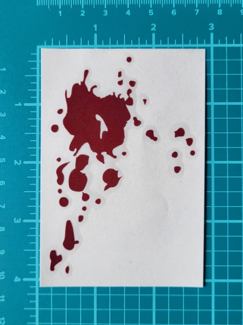 May include: A red blood splatter sticker on a white background. The sticker has a jagged, irregular shape with several smaller droplets. The sticker is placed on a blue grid background.