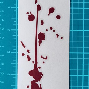 May include: A white sticker featuring a dark red splatter design. The design includes a vertical line with several splatters and droplets, creating a visual effect. The sticker is placed on a blue grid surface.