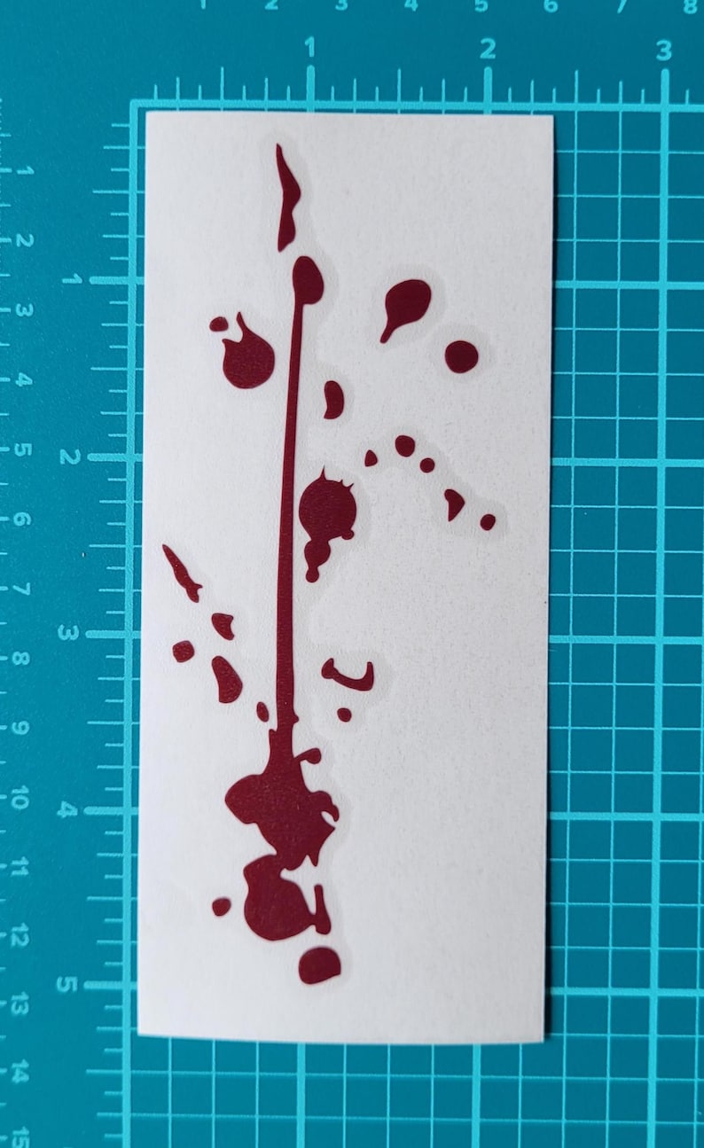 May include: A white sticker featuring a dark red splatter design. The design includes a vertical line with several splatters and droplets, creating a visual effect. The sticker is placed on a blue grid surface.