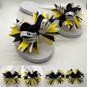 May include: Pair of white flip flops with black and yellow bee-themed bows. The bows are made with black, white, and yellow ribbon and have a glitter accent.
