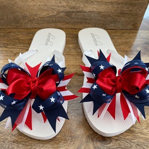 May include: A pair of white flip flops with red, white, and blue star-shaped bows. The bows are made of ribbon and mesh.