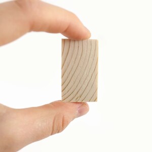 May include: A small, rectangular block of wood with a natural wood grain pattern. The block is held in a hand.