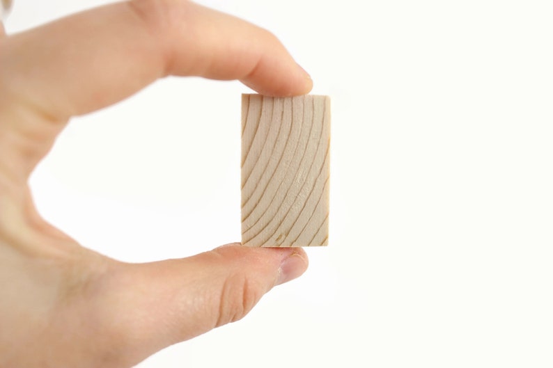 May include: A small, rectangular block of wood with a natural wood grain pattern. The block is held in a hand.