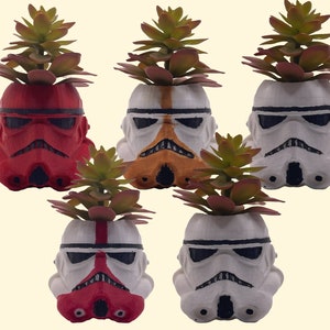 Può includere: Cinque vasi da fiori a forma di Stormtrooper di Star Wars, bianchi e rossi, con piante grasse verdi. I vasi sono dipinti in diversi schemi di colori, tra cui rosso, bianco e arancione.