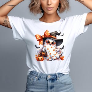 May include: White t-shirt with a Halloween graphic. The design shows a cartoon ghost wearing a witch's hat, glasses, and a polka-dot bow. The ghost is holding a pumpkin-shaped bag, with a jack-o'-lantern and bats also featured.