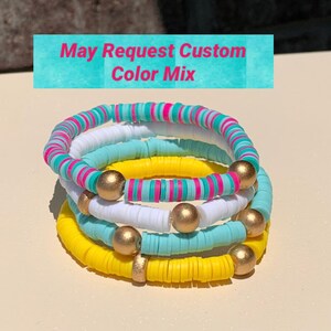 May include: A stack of four colourful bracelets made of round beads. The bracelets are yellow, white, light blue, and pink and white striped. Each bracelet has gold beads interspersed throughout.