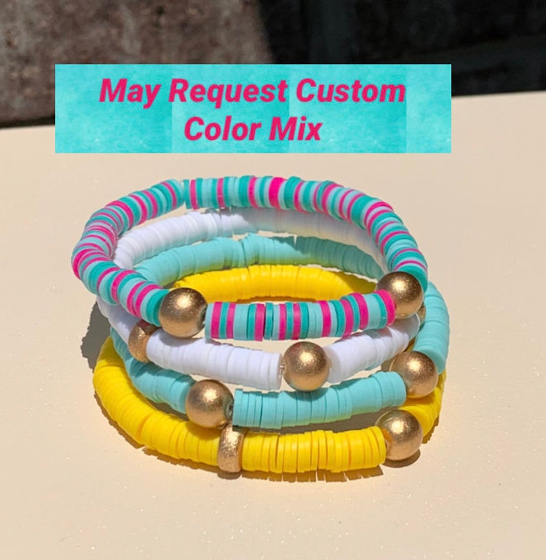 May include: A stack of four colourful bracelets made of round beads. The bracelets are yellow, white, light blue, and pink and white striped. Each bracelet has gold beads interspersed throughout.