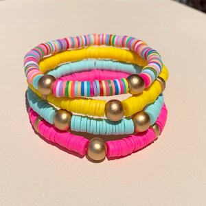 May include: A stack of four colourful bracelets made of small, round beads. The bracelets are pink, yellow, light blue, and multi-coloured. Each bracelet has gold beads interspersed throughout.
