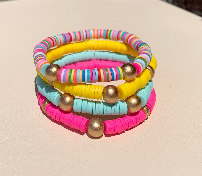 May include: A stack of four colourful bracelets made of small, round beads. The bracelets are pink, yellow, light blue, and multi-coloured. Each bracelet has gold beads interspersed throughout.