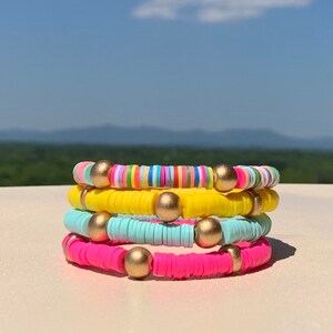 May include: Four colourful bracelets stacked on top of each other. The top bracelet is rainbow striped, the second is yellow, the third is light blue, and the bottom bracelet is pink. Each bracelet has gold beads.