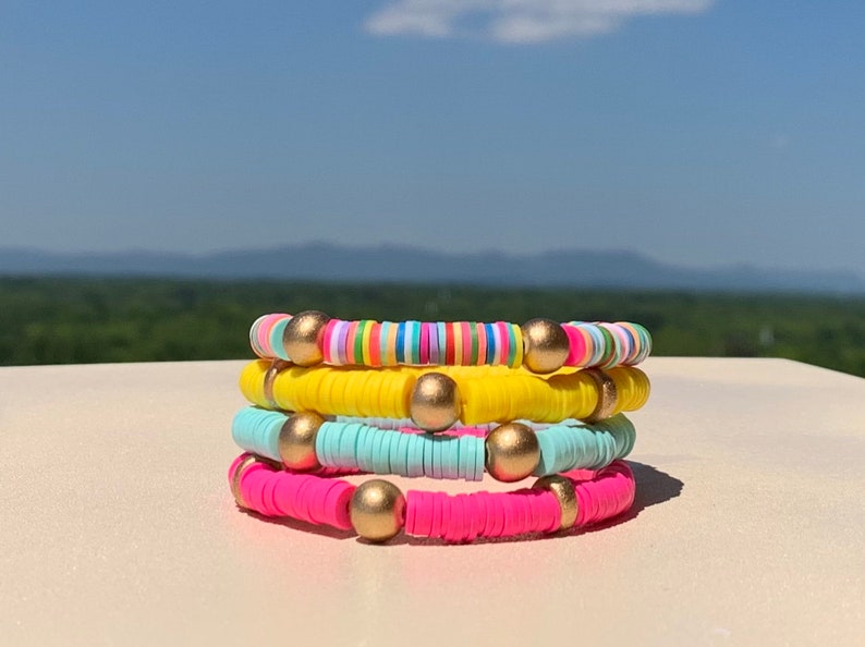 May include: Four colourful bracelets stacked on top of each other. The top bracelet is rainbow striped, the second is yellow, the third is light blue, and the bottom bracelet is pink. Each bracelet has gold beads.