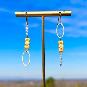 May include: A pair of gold-toned earrings with a unique design. Each earring features an oval-shaped hoop, three small, light-brown beads, and a decorative chain or beaded accent. The earrings are displayed against a bright blue sky background.
