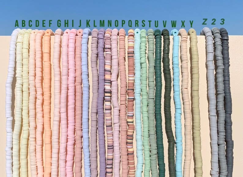 May include: A collection of 26 strands of colourful polymer clay beads, labelled with letters A through Z, and two additional strands labelled 2 and 3. The beads are all round and flat, and they are arranged in a row against a white background.