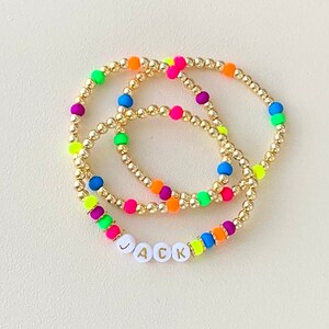 May include: Three gold beaded bracelets with neon-colored beads in pink, orange, yellow, green, blue, and purple. One bracelet spells out the name "JACK" in white letter beads.