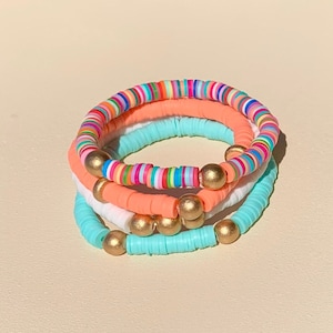 May include: A set of three colourful beaded bracelets. The bracelets are made of small, round beads in shades of pink, blue, white, and rainbow. The bracelets are accented with gold beads.
