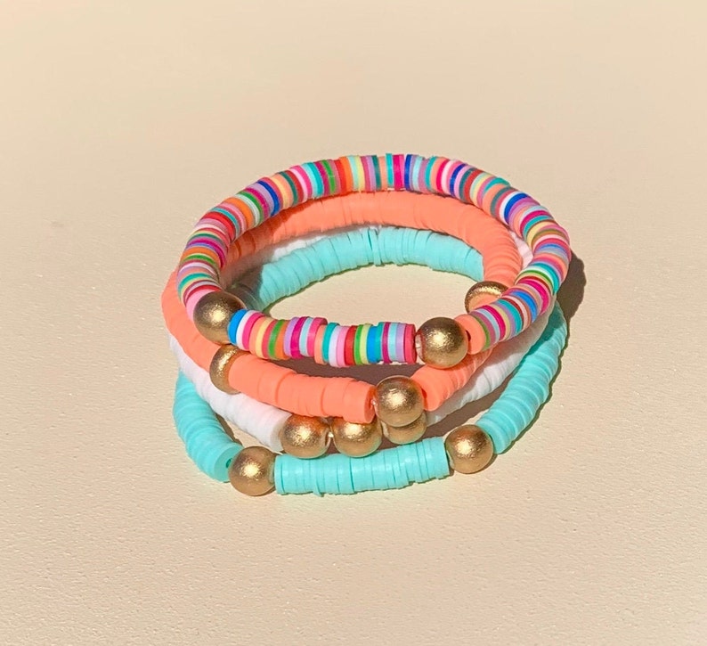 May include: A set of three colourful beaded bracelets. The bracelets are made of small, round beads in shades of pink, blue, white, and rainbow. The bracelets are accented with gold beads.