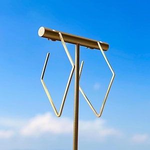 May include: Gold-colored geometric earrings with a diamond shape, displayed on a matching gold stand. The earrings are suspended against a clear, bright blue sky, creating a striking contrast.