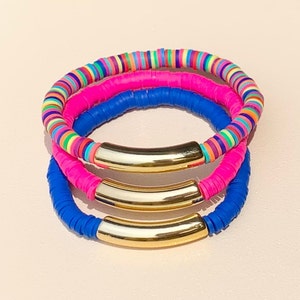 May include: Three colorful bracelets with gold metal accents. The bracelets are stacked on top of each other. The top bracelet is multi-colored, the middle bracelet is pink, and the bottom bracelet is blue.
