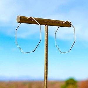 May include: A pair of silver octagon hoop earrings on a gold display stand against a blue sky background.