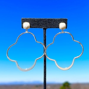 May include: Silver earrings with a cloud shape and a white pearl accent. The earrings are hanging from a black display stand against a blue sky background.