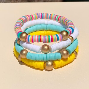 May include: A stack of four colourful bracelets with gold beads. The bracelets are made of small, round, flat beads in shades of yellow, blue, white, and rainbow.