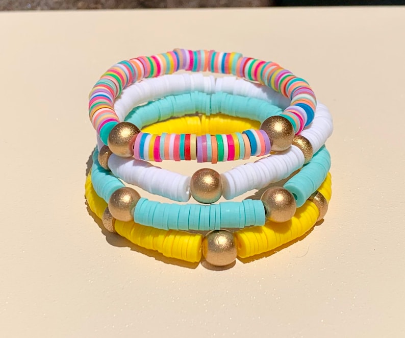 May include: A stack of four colourful bracelets with gold beads. The bracelets are made of small, round, flat beads in shades of yellow, blue, white, and rainbow.