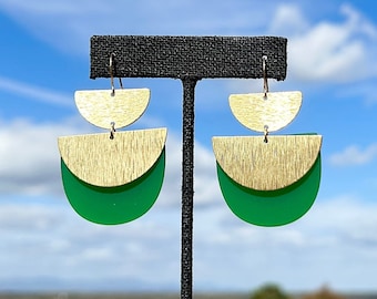 May include: Geometric earrings with a layered design. The earrings feature a gold-colored half-circle on top of a gold-colored half-circle, which is on top of a green half-circle. The earrings are hanging from a gold-colored hook.