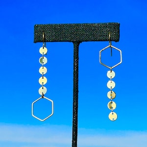 May include: Gold tone earrings with a hexagonal shape and a chain of round discs. The earrings are displayed on a black stand against a blue sky background.