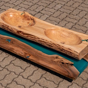 K&ouml;nnte beinhalten: Ein Doppelwaschtisch aus einer gro&szlig;en, nat&uuml;rlichen Holzplatte mit zwei ausgeh&ouml;hlten Becken. Das Holz hat eine nat&uuml;rliche, rustikale Oberfl&auml;che und steht auf einem Sockel aus dem gleichen Holz mit einer Einlage aus tealblauem Epoxidharz.