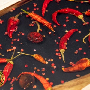 May include: A close-up of a wooden tray with red chili peppers and pink peppercorns embedded in clear resin. The tray has a dark background and a wooden border.