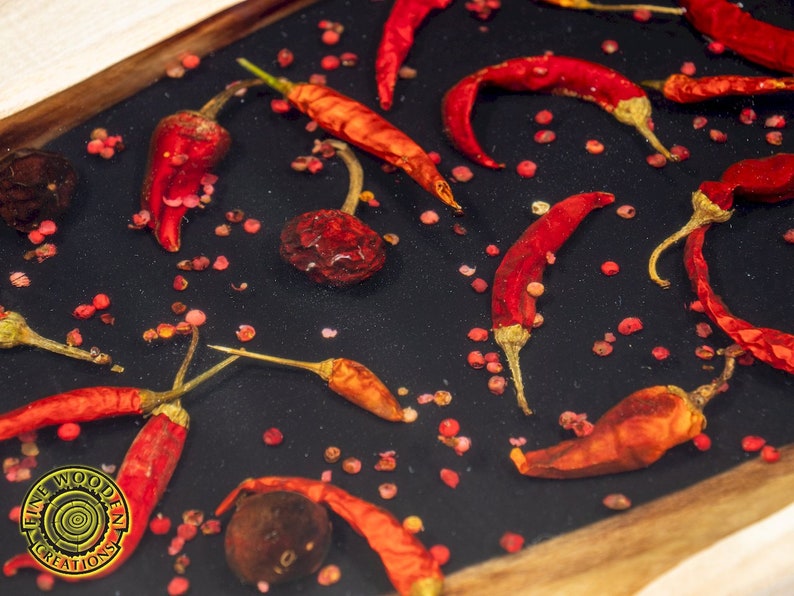 May include: A close-up of a wooden tray with red chili peppers and pink peppercorns embedded in clear resin. The tray has a dark background and a wooden border.