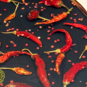 May include: A close-up of a black resin tray with red chili peppers and pink peppercorns. The tray is round and has a wooden edge.