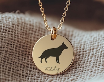 May include: A gold-colored necklace with a circular pendant featuring a black silhouette of a dog. The pendant also has the name "Teddy" engraved in script. The necklace is displayed on a textured, beige fabric.