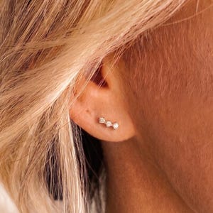 May include: A silver-toned ear climber earring featuring three small, clear, round-cut stones arranged in a linear, ascending pattern. The earring is secured to the earlobe, following the natural curve of the cartilage with a delicate, minimalist design.