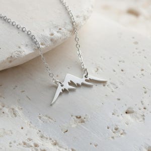 May include: A silver-colored necklace featuring a mountain range pendant. The pendant is a minimalist design with sharp peaks and a delicate chain. The necklace is displayed on a textured, off-white surface.