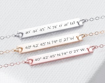 May include: Three gold bar necklaces with a heart charm at the top. Each necklace has a different color chain: silver, gold, and rose gold. The bars are engraved with the coordinates "40° 42′ 45° N 74° 0′ 21" W."